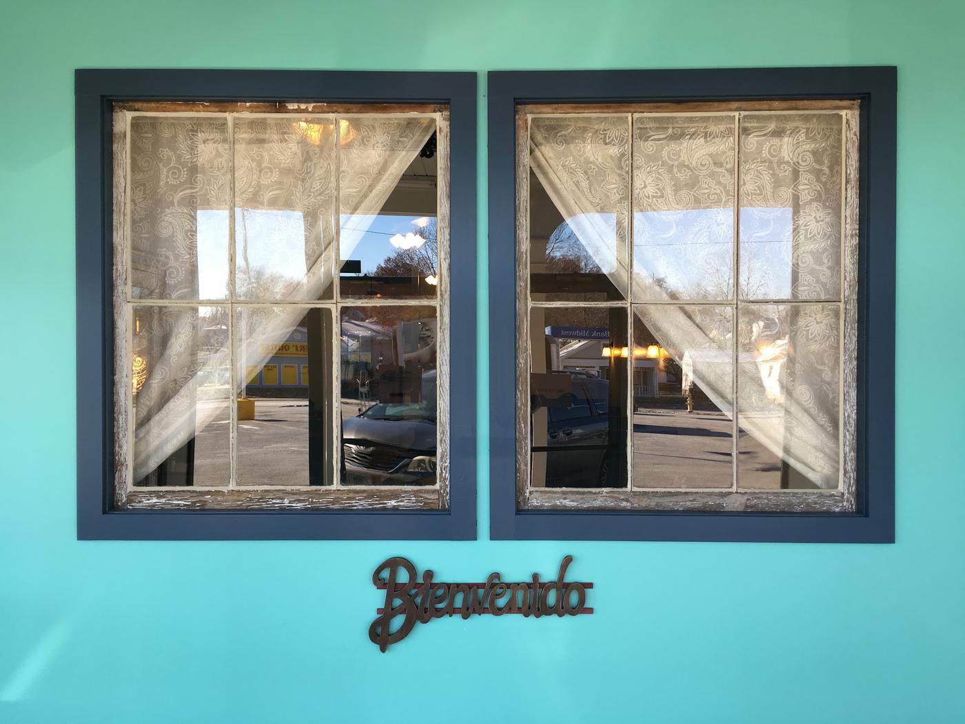 Two salvaged sash windows with lace curtains mounted on a turquoise wall, with a cut-out Bienvenido sign below them