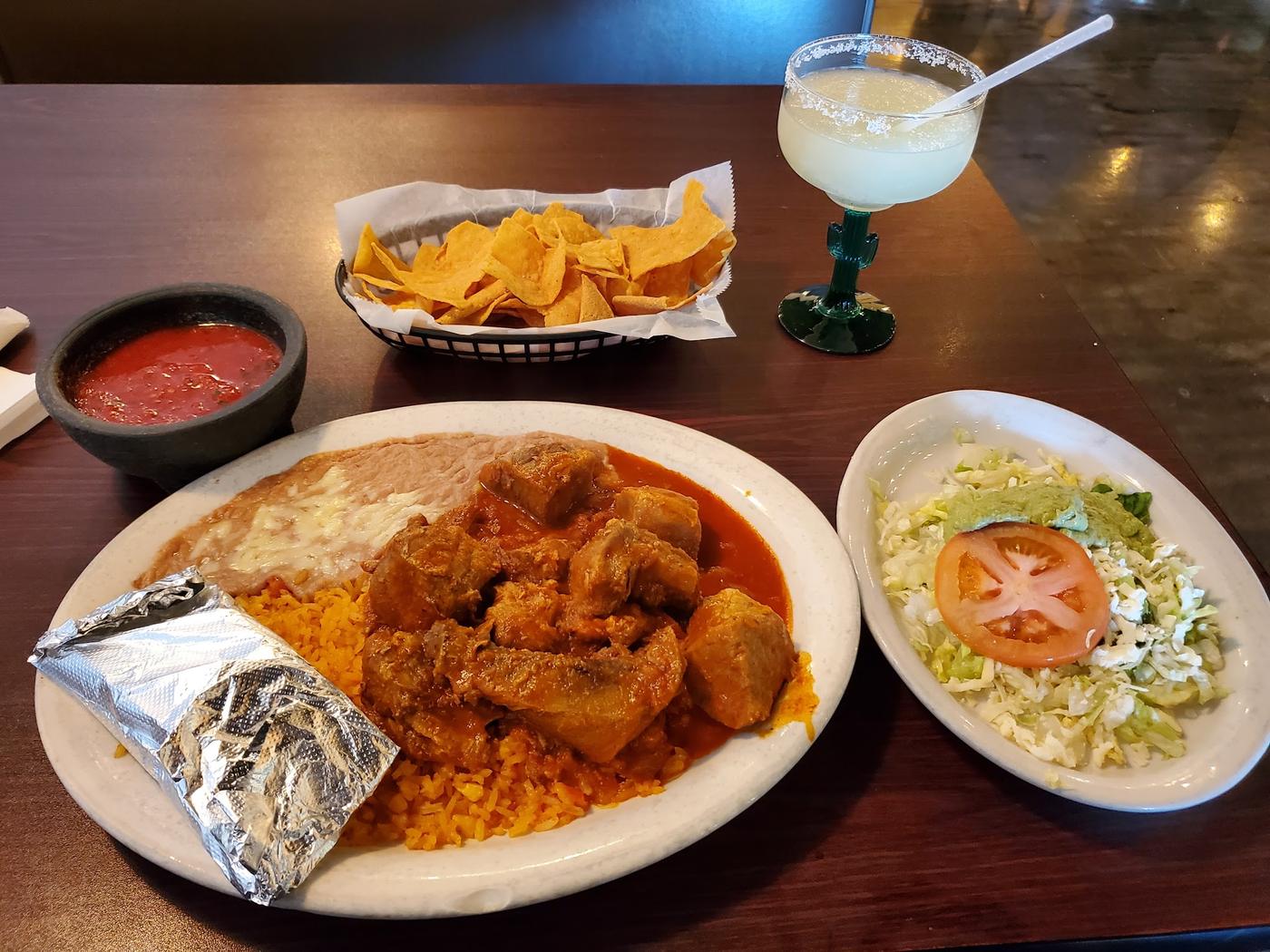 A plate of chicken in red chile sauce with yellow rice and beans, beside chips, salsa and a frozen margarita